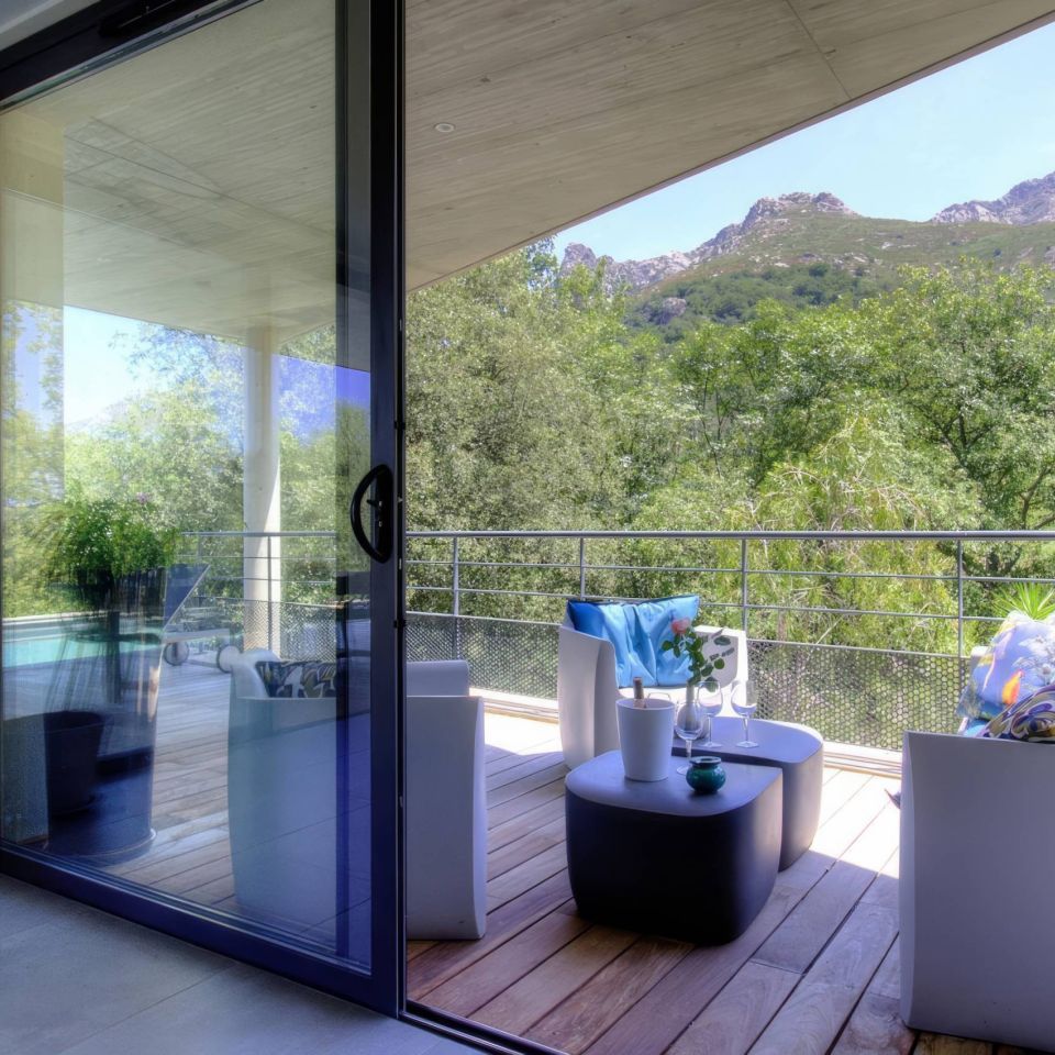 A modern balcony with furniture overlooks a lush, green landscape and mountains under a clear blue sky.