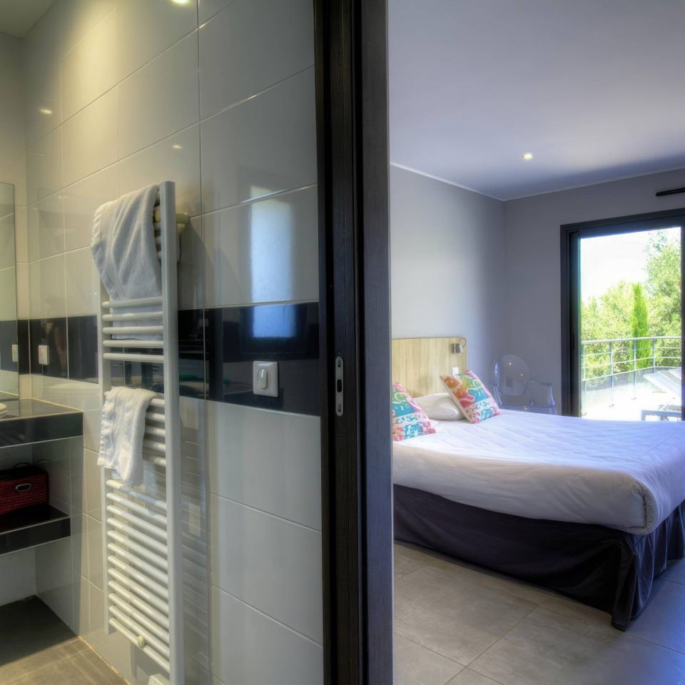 Modern bedroom and bathroom with a sleek design, including a bed, mirror, and view of greenery outside.