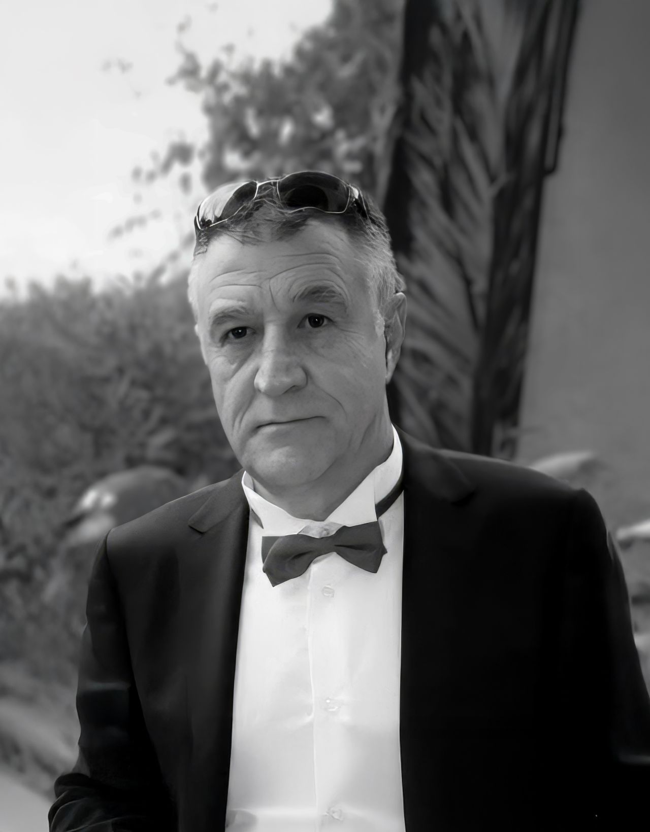 Un homme en smoking avec un nœud papillon et des lunettes de soleil, se tenant à l'extérieur dans un arrière-plan doucement flou.