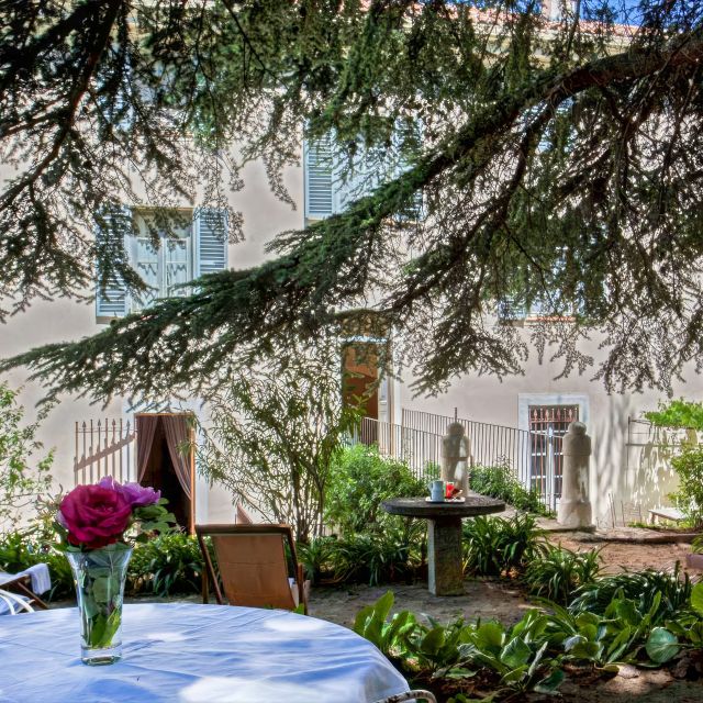 A serene garden scene featuring a table with a flower vase, greenery, and a glimpse of a house in the background.