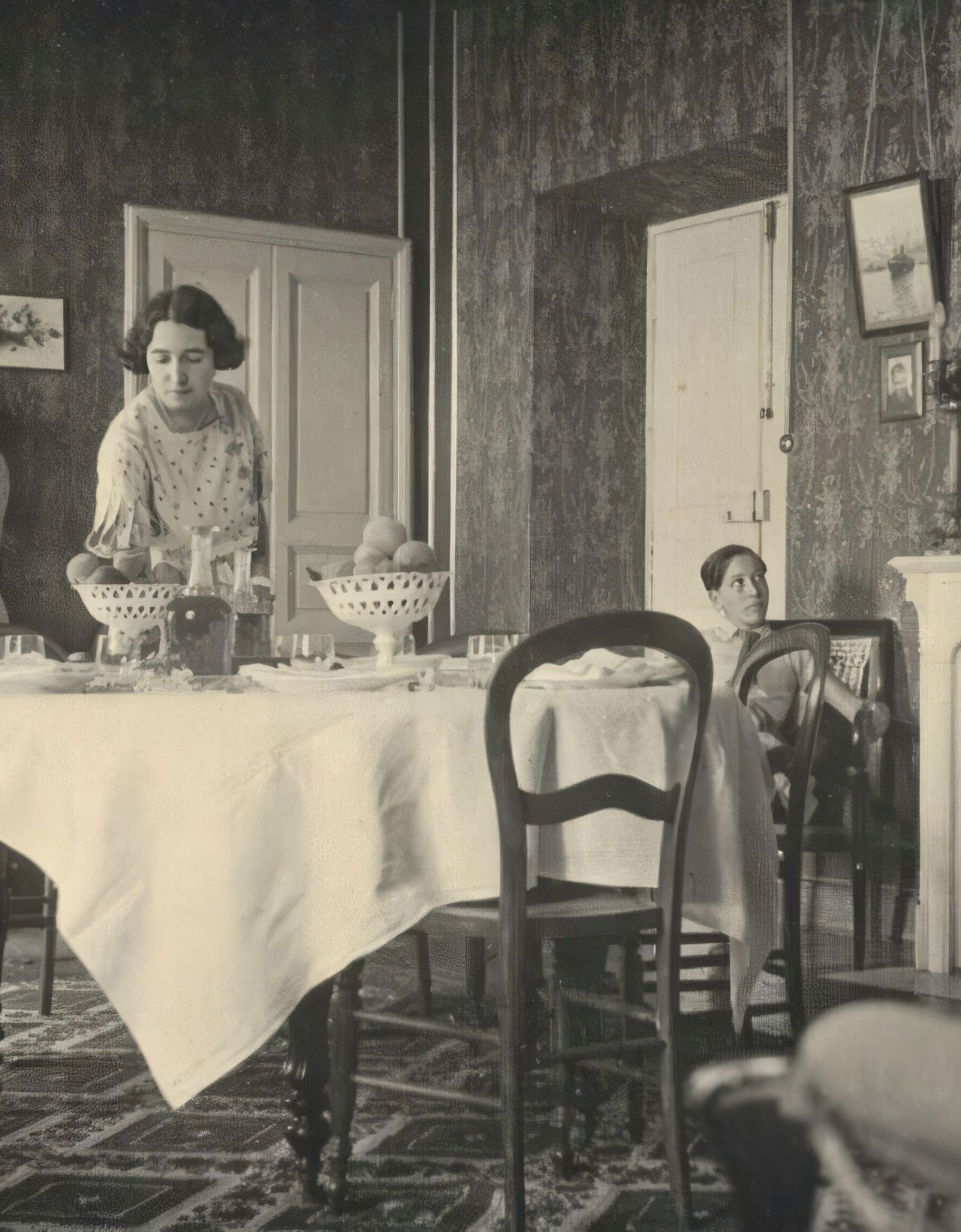 A vintage kitchen scene featuring three women, a dining table set for a meal, and ornate wallpaper, capturing everyday life.