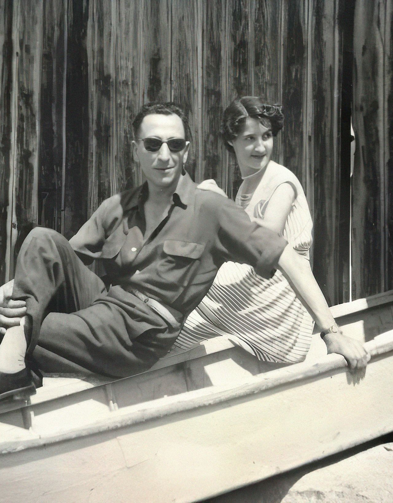A couple sits playfully in a small boat, smiling, against a rustic wooden backdrop.