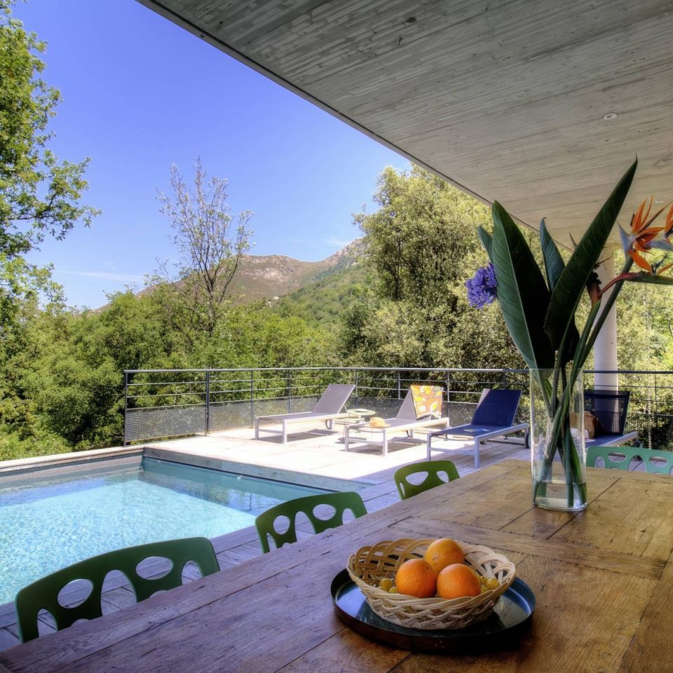 Ensoleillé patio avec piscine, table à manger, fleurs vives et sièges, entouré de verdure luxuriante et de montagnes.