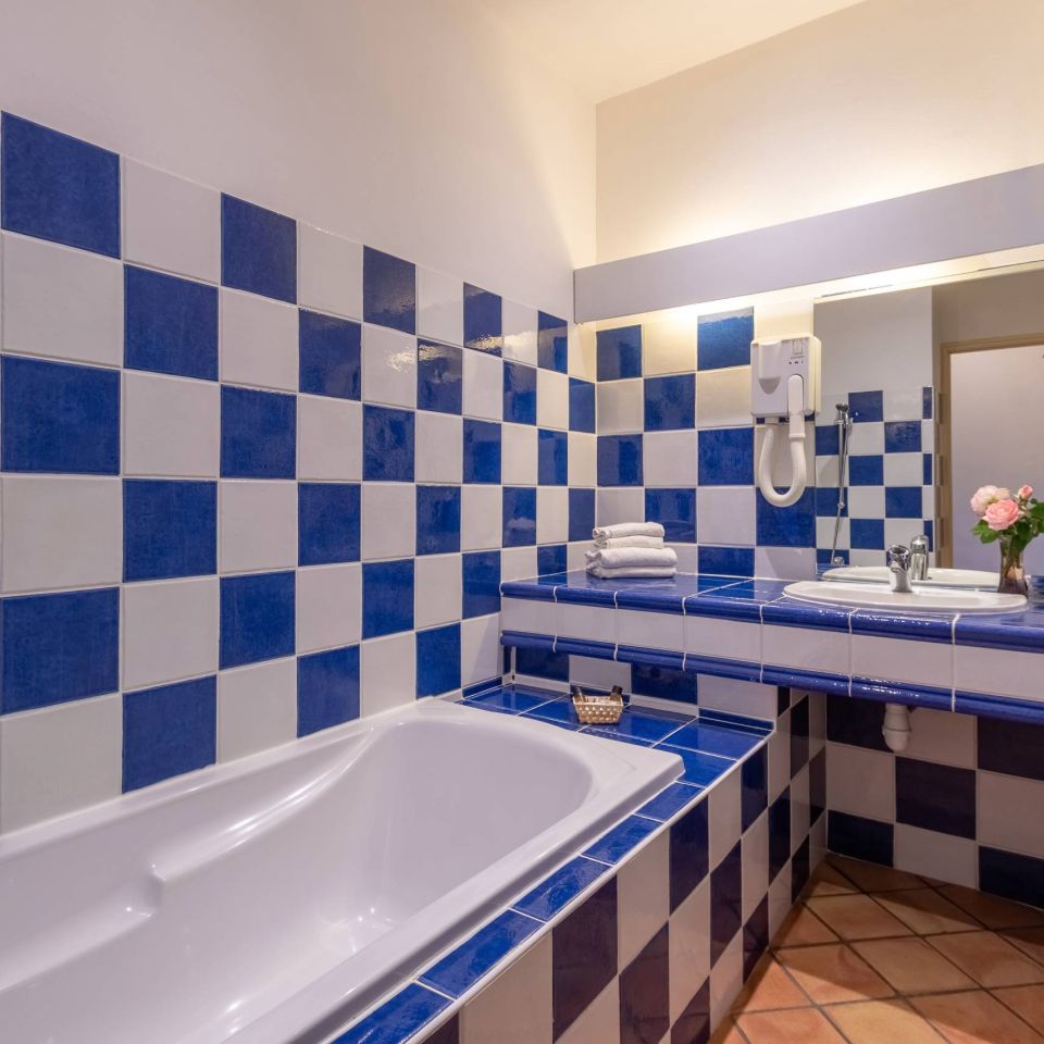 Ein helles Badezimmer verfügt über blau-weiße kariertes Fliesen, eine Badewanne, ein Waschbecken und eine kleine Vase mit Blumen.