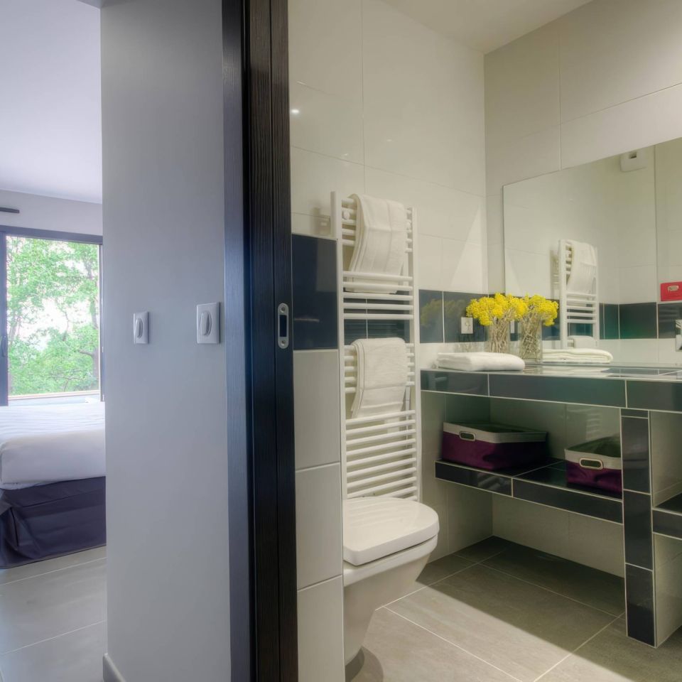 Modern bathroom with towels, flowers, and a view towards a bedroom and outdoor area with greenery.