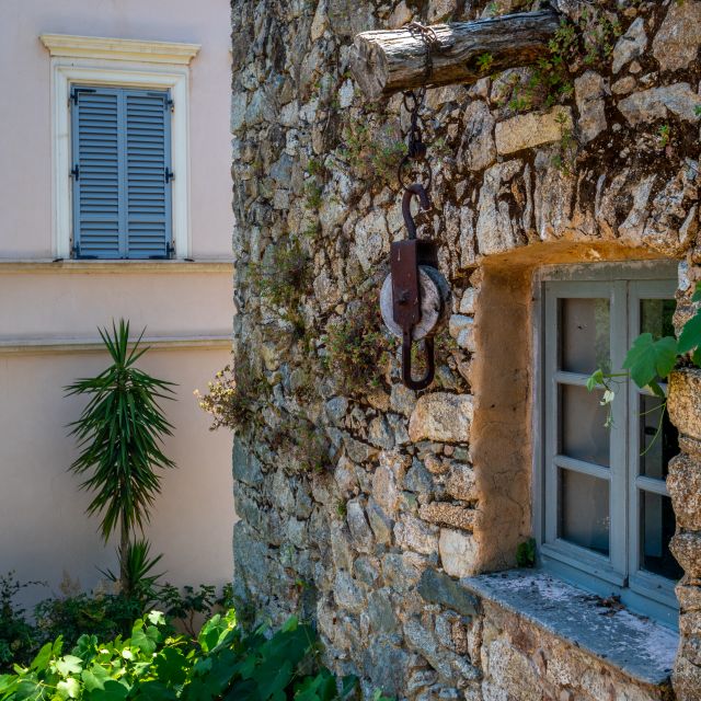 Eine Steinmauer mit einer Rolle, grauem Fenster und Grünpflanzen, vor einem Pastellgebäude mit blauen Fensterläden.