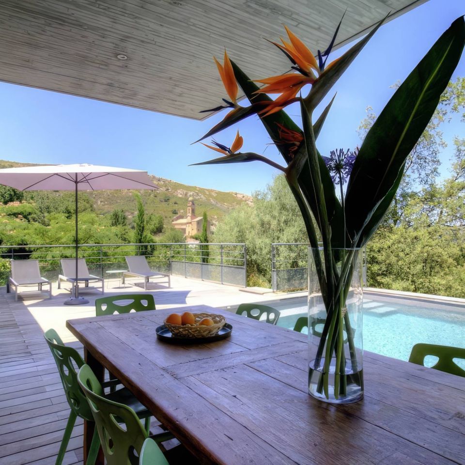 A sunny terrace with a wooden table, green chairs, and a swimming pool surrounded by lush greenery and mountains.