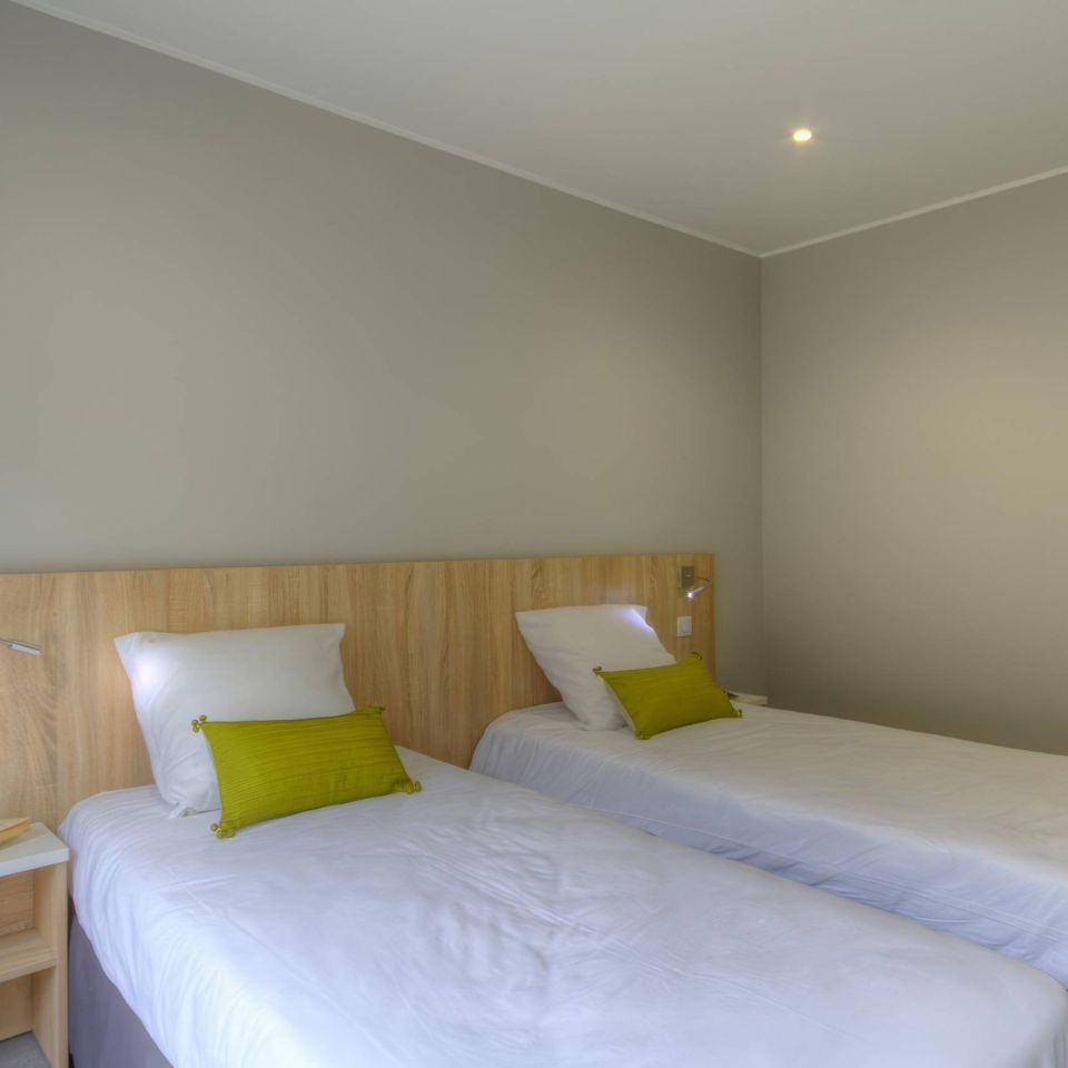 A simple, modern room featuring two single beds with white linens and green decorative cushions against a grey wall.