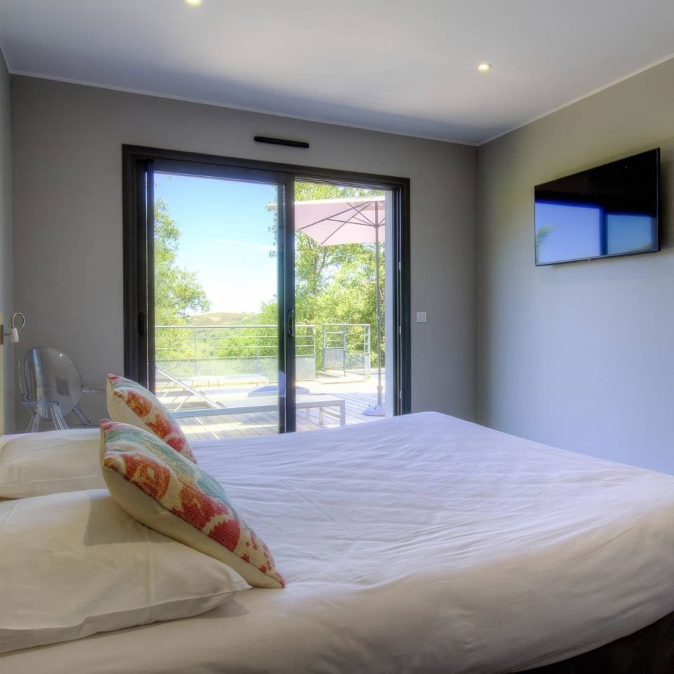 Modern room with a large bed, colourful cushions, TV, and sliding doors opening to a sunny outdoor area.
