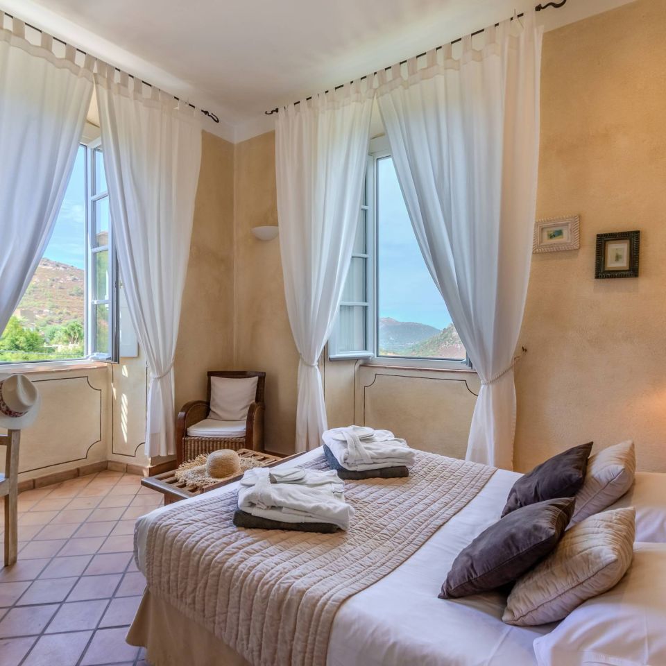 Camera luminosa e serena con un letto accogliente, grandi finestre e tende morbide, che mostrano una vista panoramica e un arredamento raffinato.