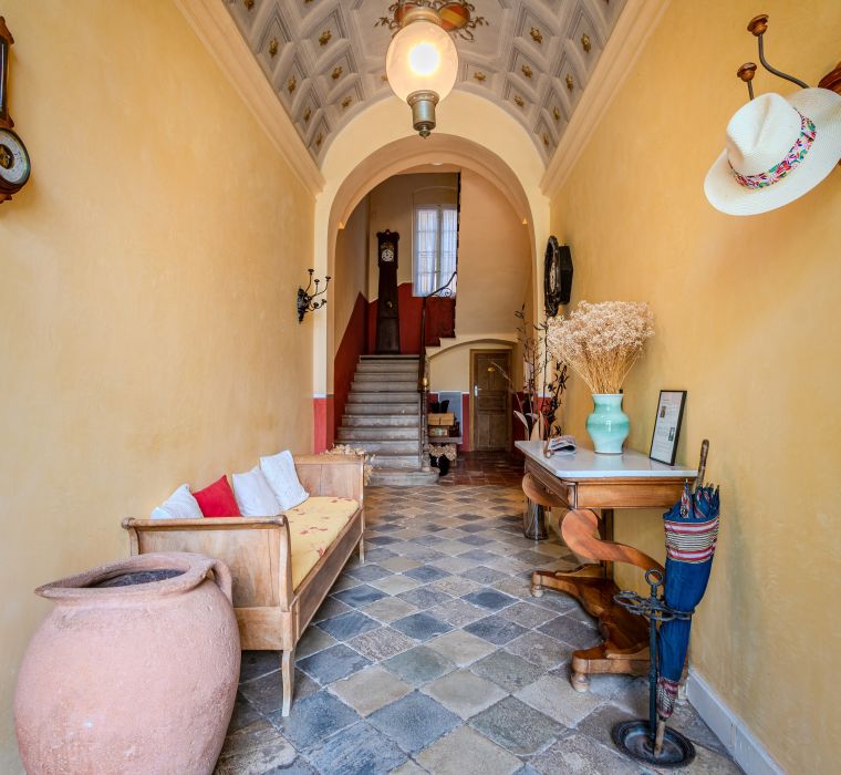 Un couloir accueillant présente un escalier, un sol à motifs, un banc, des décorations et de la lumière naturelle. Ambiance chaleureuse et accueillante.