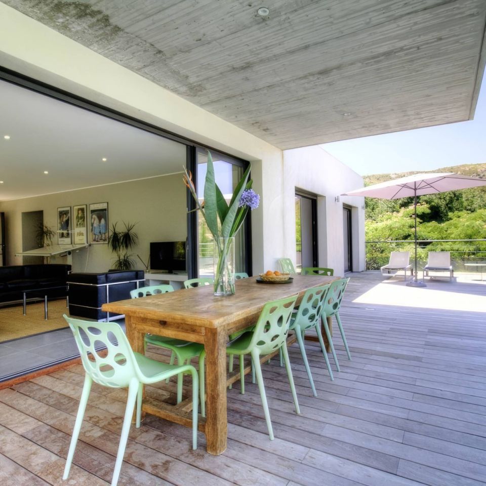 Modern hotel patio with a wooden table, green chairs, and a view of nature, featuring an open interior and pool area.