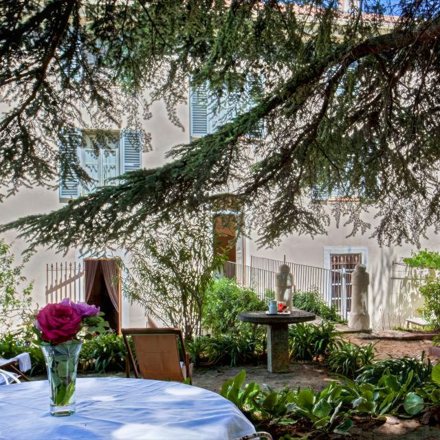 Una scena serena di giardino con un tavolo e un vaso di fiori, verdeggianti e un accenno di una casa sullo sfondo.
