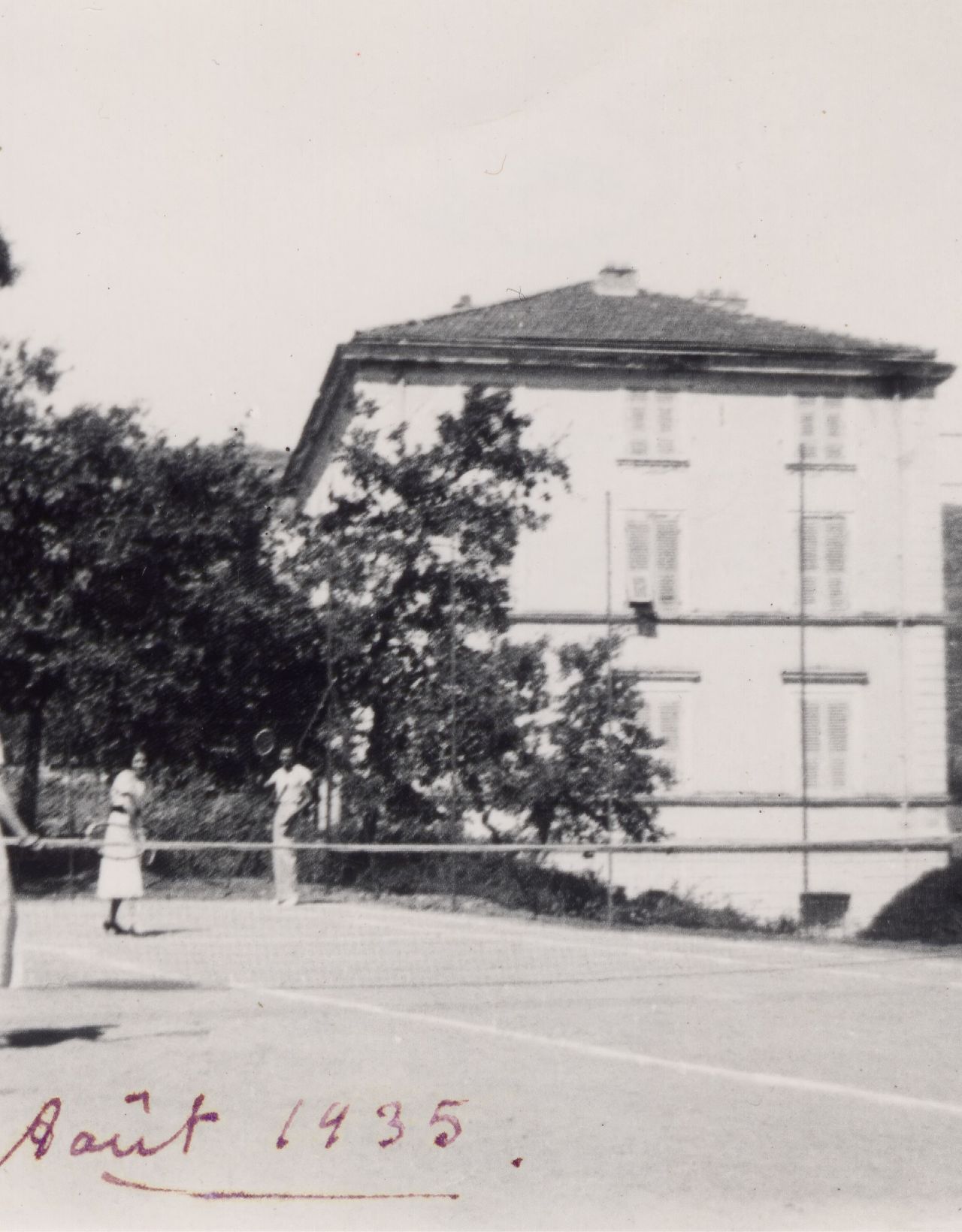 Ein historisches Schwarz-Weiß-Foto von vier Personen, die Tennis in der Nähe eines Hauses spielen, datiert auf August 1935.