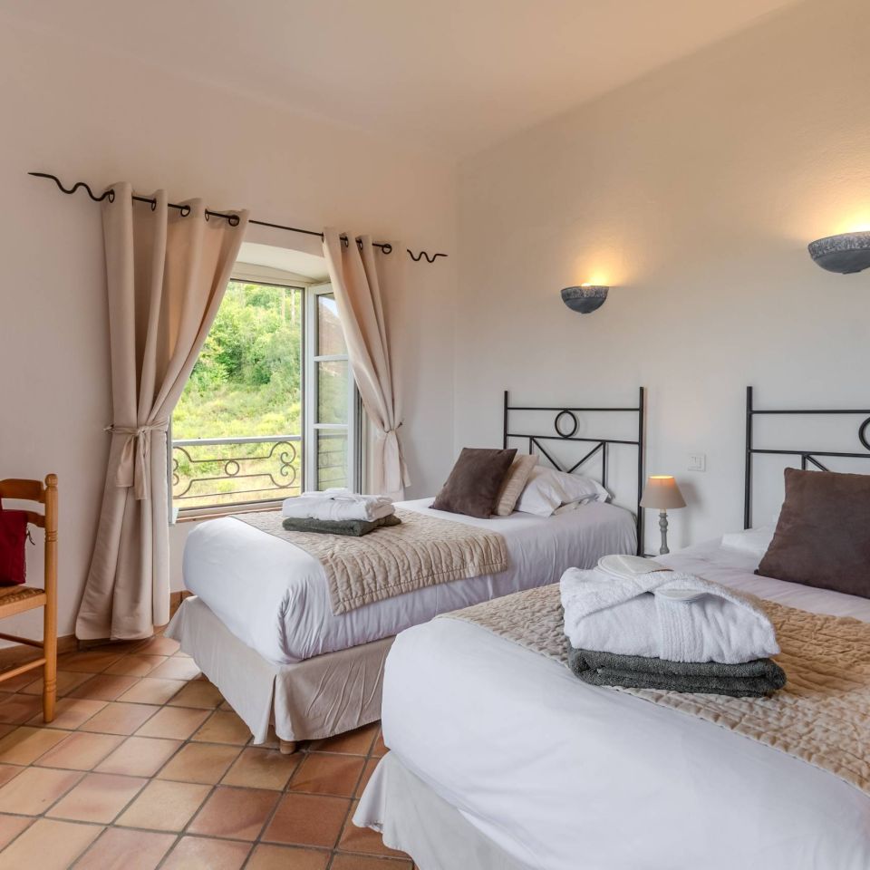A cosy bedroom with two beds, warm lighting, a view of nature, and a wooden chair and table.