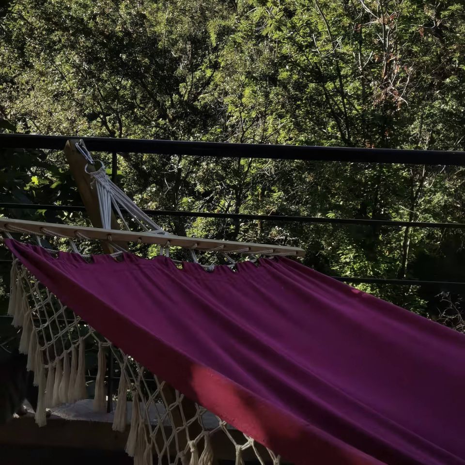 A purple hammock hangs on a balcony, surrounded by lush green trees and mountainous scenery under a blue sky.