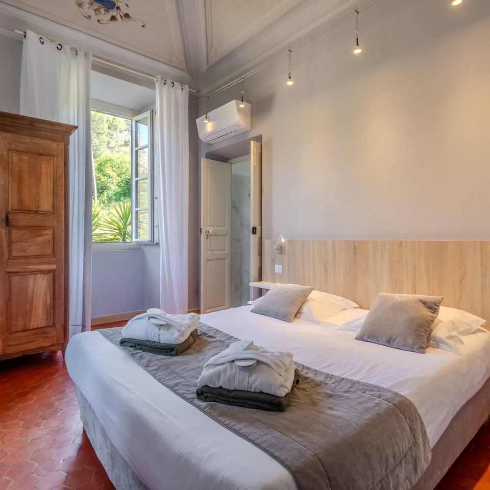 Bright bedroom with a wooden wardrobe, large window, and neatly made bed featuring soft pillows and towels.