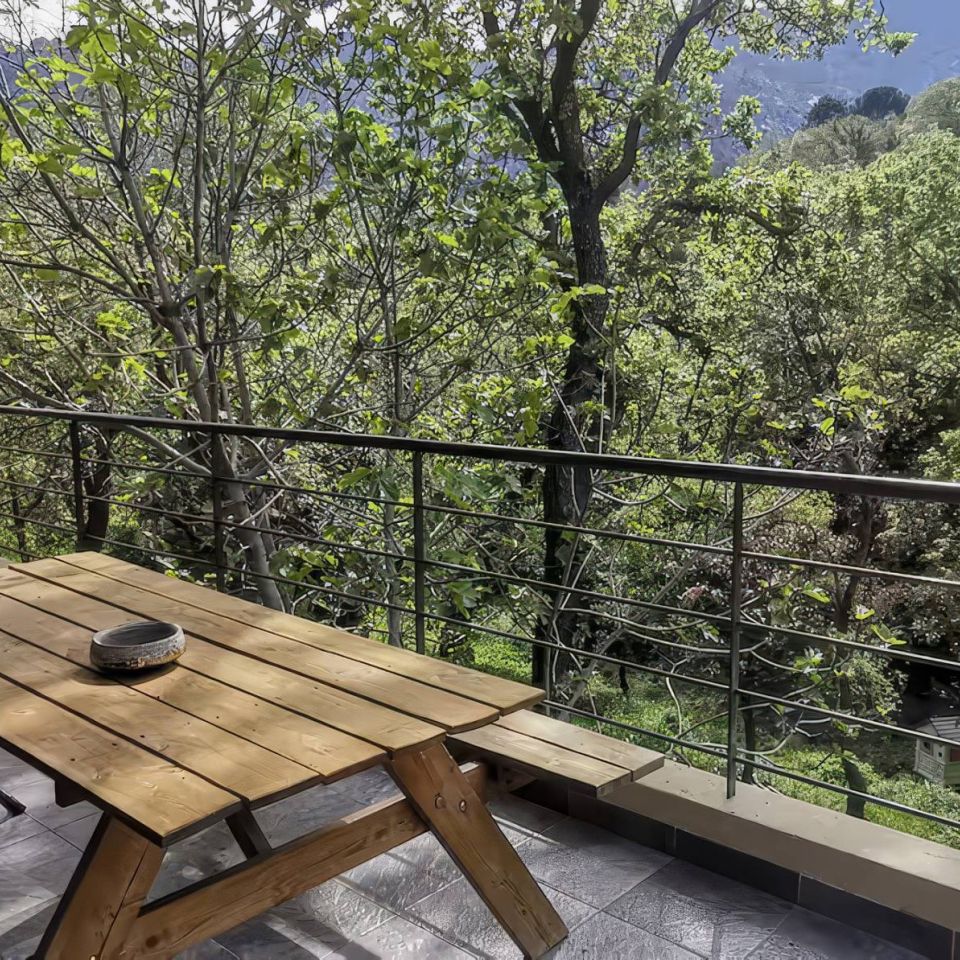 A wooden table and chairs on a balcony surrounded by lush greenery and distant mountains in a serene outdoor setting.
