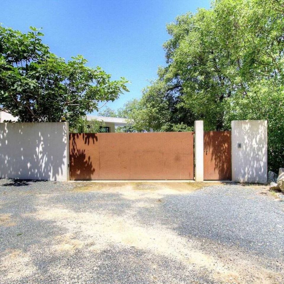 A rust-coloured gate flanked by white walls, surrounded by lush greenery under a clear blue sky.
