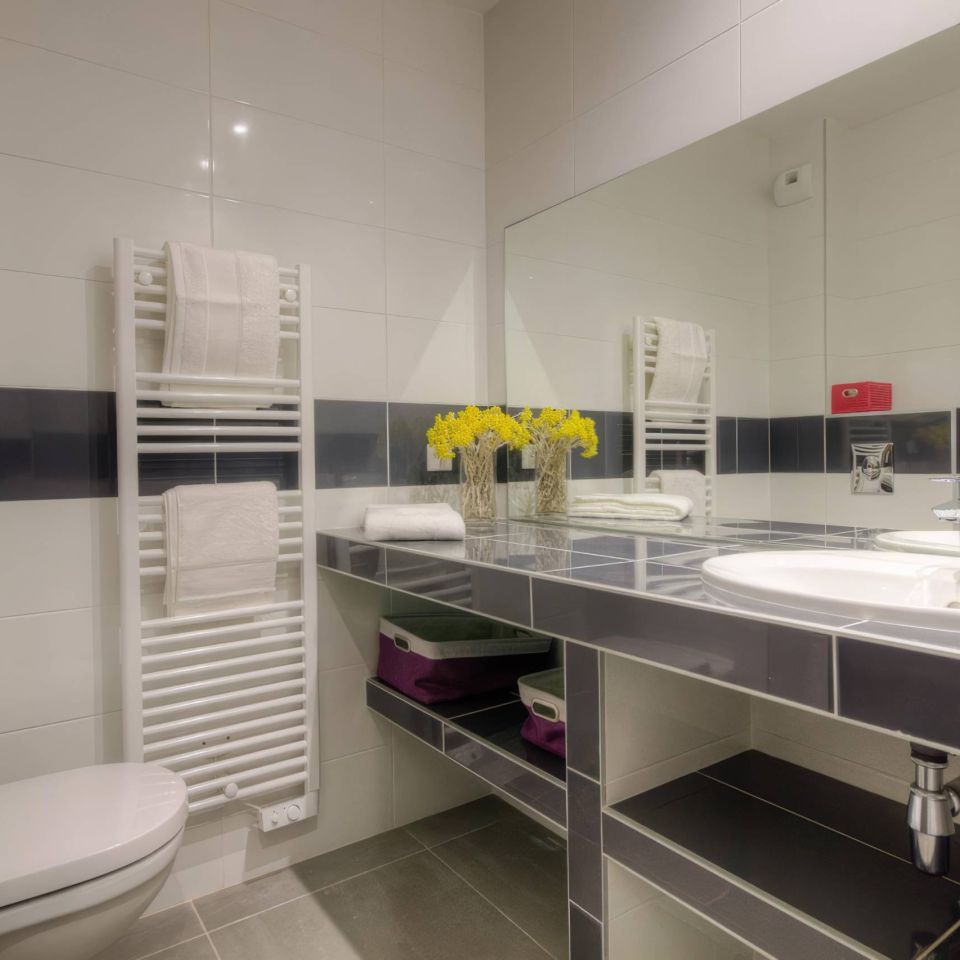 Modern bathroom featuring a toilet, sink, towel rack, and flowers, with sleek tiles and a mirror.