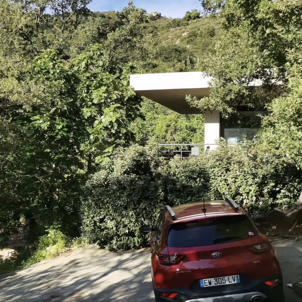 Une voiture rouge garée sur un chemin en gravier entouré d'arbres verdoyants et d'un bâtiment moderne en arrière-plan.