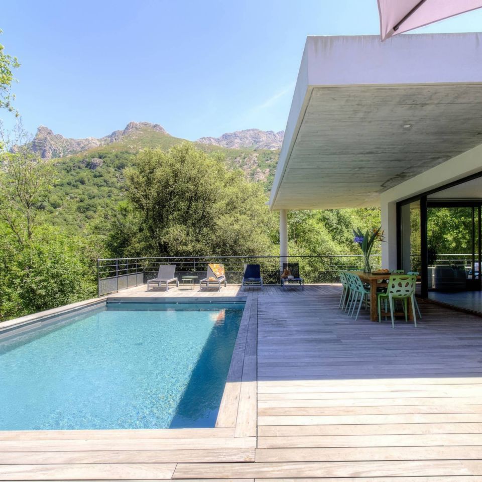 Modern house with a pool and deck, surrounded by lush greenery and mountains under a clear blue sky.