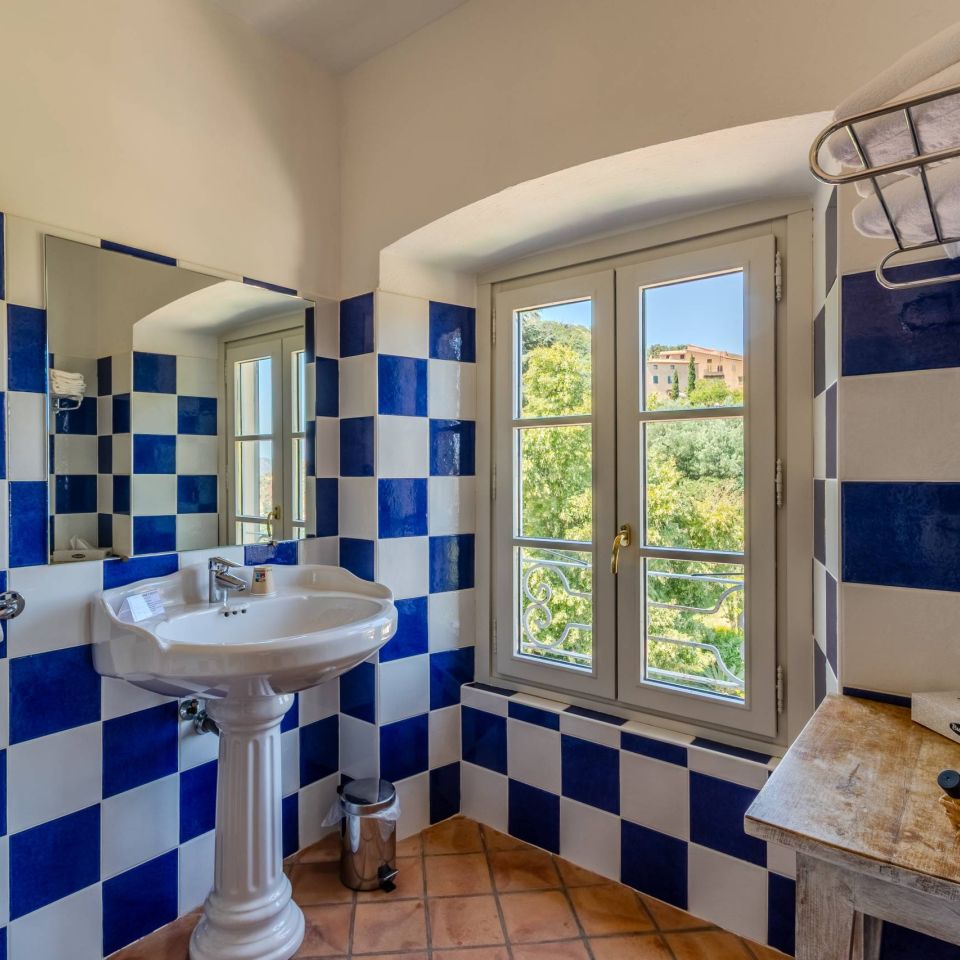 Bright bathroom with blue and white tiles, sink, mirror, and a window overlooking greenery and buildings outside.