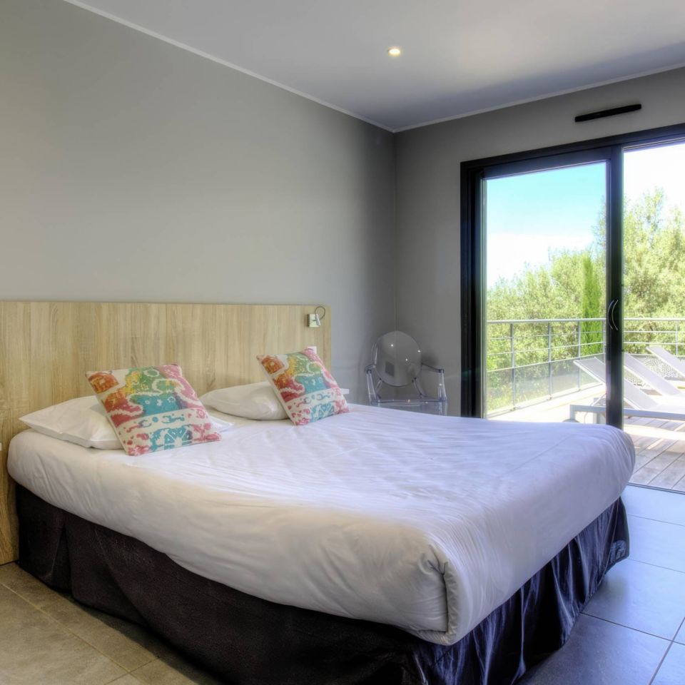 Modern chambre avec un lit double, des coussins colorés et une grande fenêtre donnant sur un balcon et la verdure à l'extérieur.