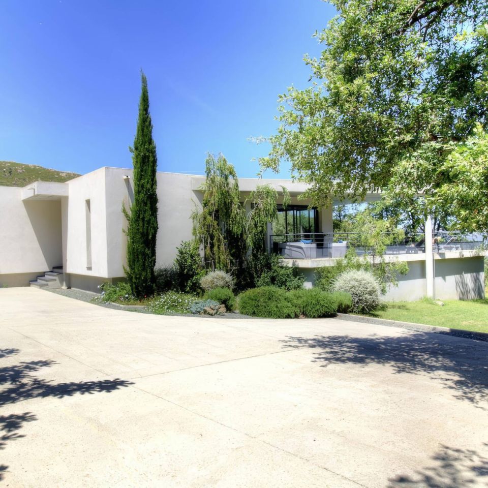 Modern house with minimalist design, surrounded by greenery and a clear blue sky. Driveway leading to the entrance.