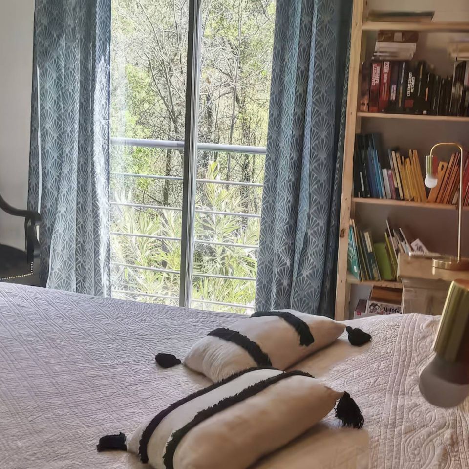Cozy bedroom with a bed, plush pillows, a chair, bookshelf, and large windows showcasing greenery outside.