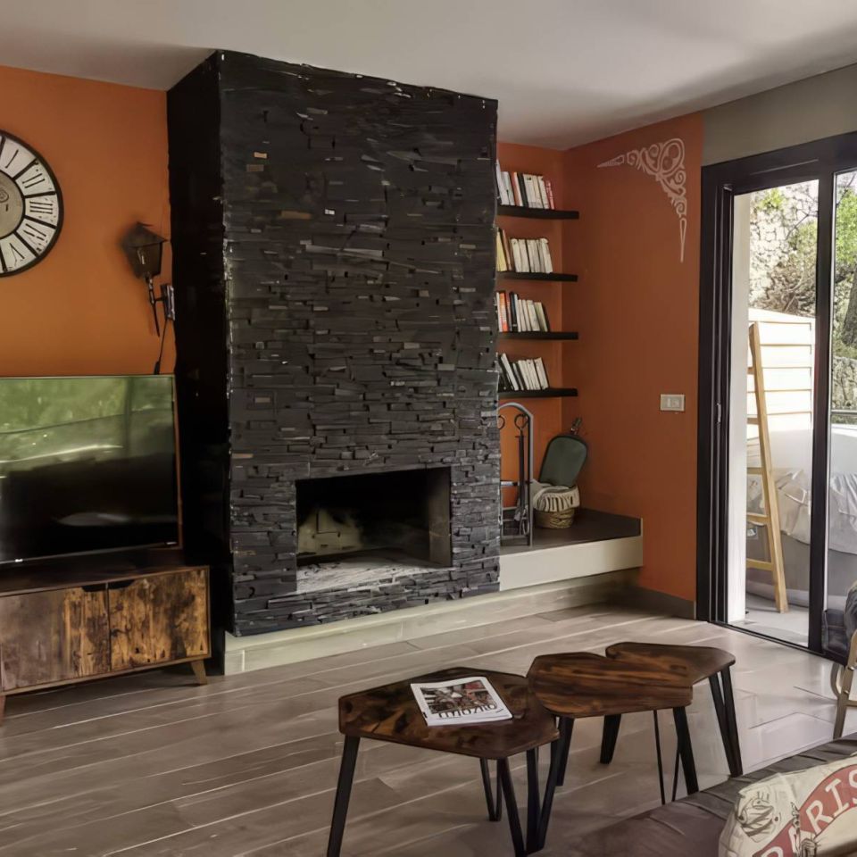 A cosy living room with a stone fireplace, TV, bookshelves, and large windows showcasing greenery outside.