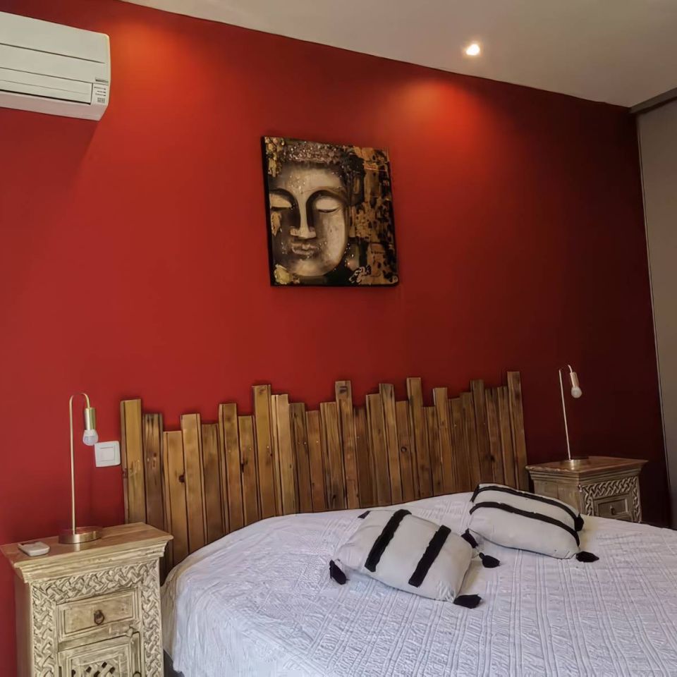 A cosy bedroom with red walls, a wooden bed head, Buddhist art, and bookshelves. Stylish and inviting décor.