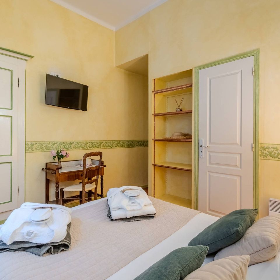 Cosy bedroom with yellow walls, a bed with towels, a small table, and a TV mounted on the wall.