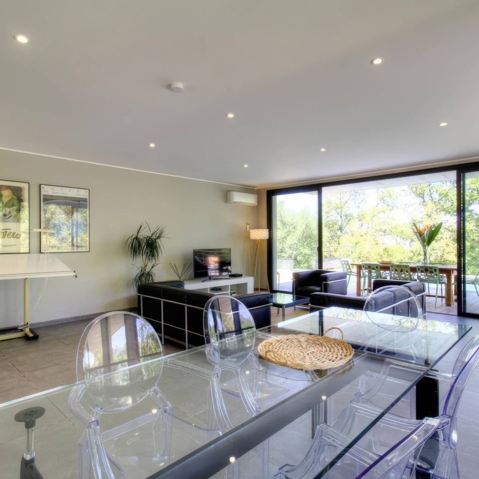 Modern living space with a glass dining table, sleek furniture, and large windows showcasing greenery.