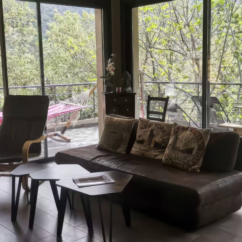 Modern living room with a sofa, cosy chairs, and a balcony view of lush greenery and a hammock outside.