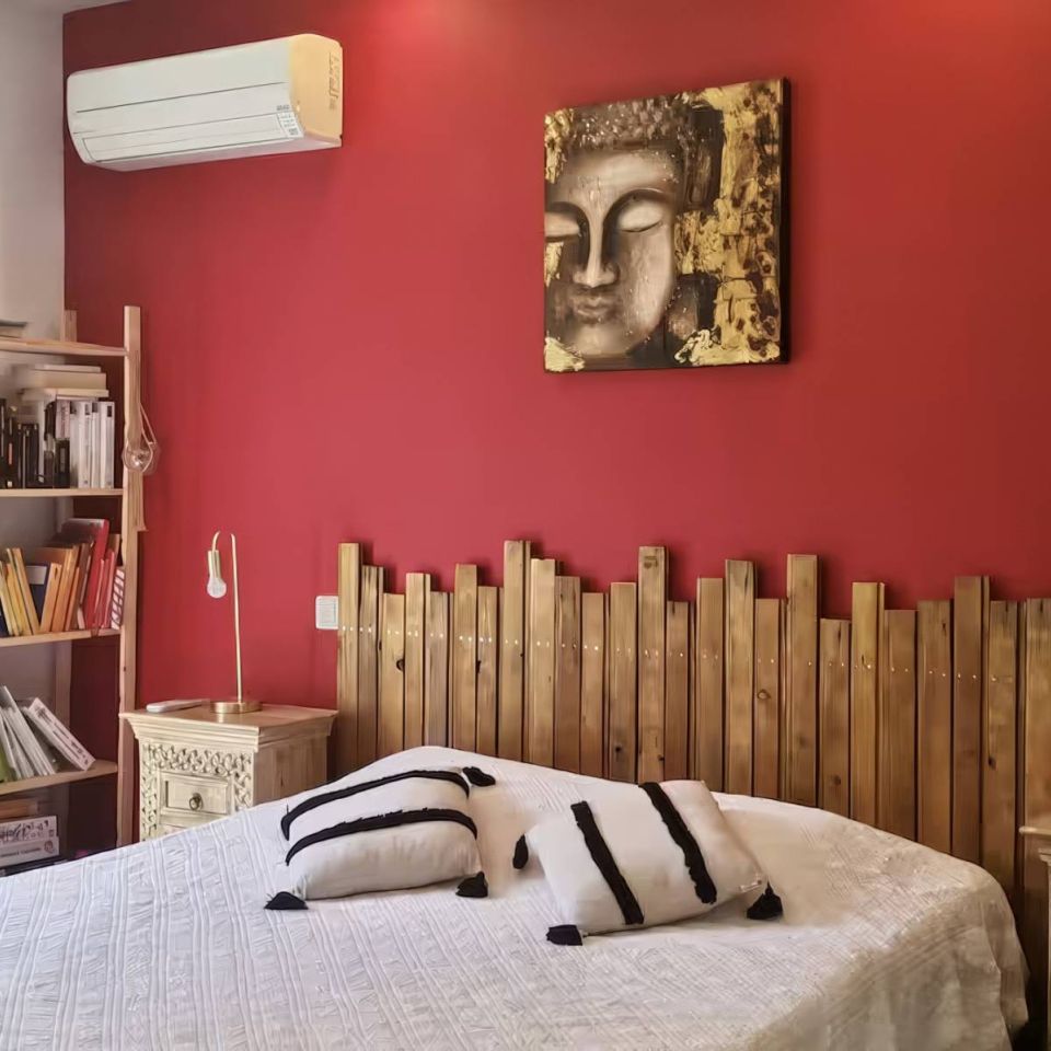 Cozy bedroom with red walls, wooden accents, a bed with patterned pillows, and a bookshelf beside a window.