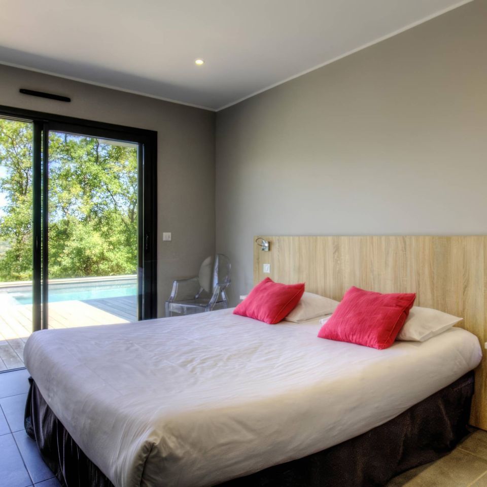 Modern room featuring a large bed with red pillows, glass doors, and a view of a pool and nature outside.