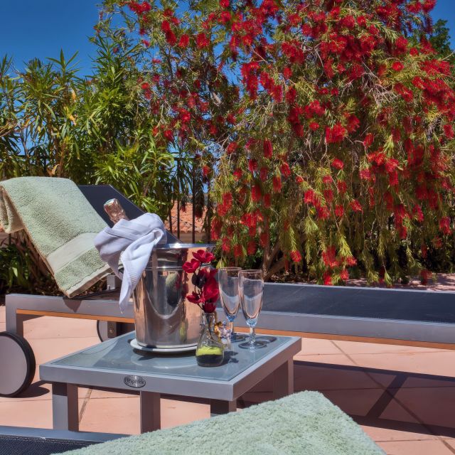 Un patio sereno con una sdraio, un asciugamano verde, champagne, flûte e fiori rossi vivaci sullo sfondo.