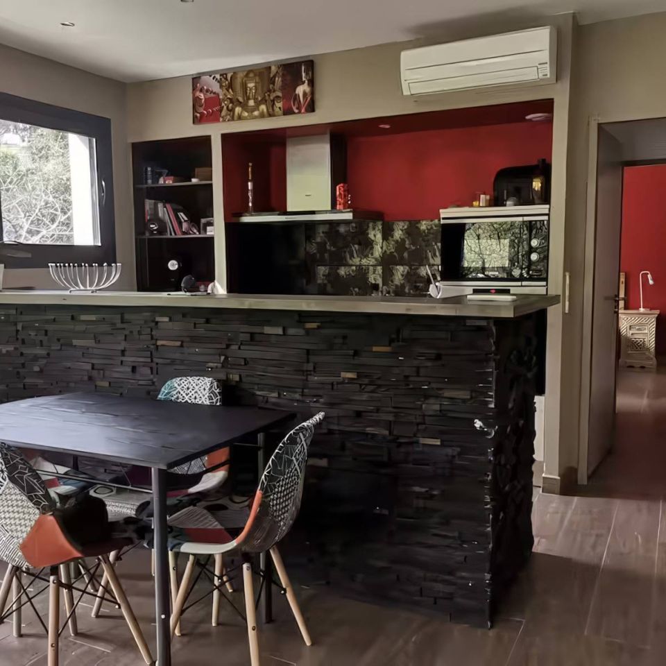 Modern kitchen with a stone bar, colourful chairs, and a view of greenery through large windows.