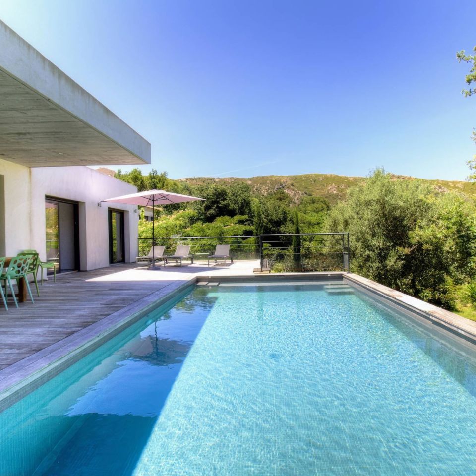Maison moderne avec piscine, chaises vertes et vue sur les collines sous un ciel bleu clair. Parfaite pour la détente.