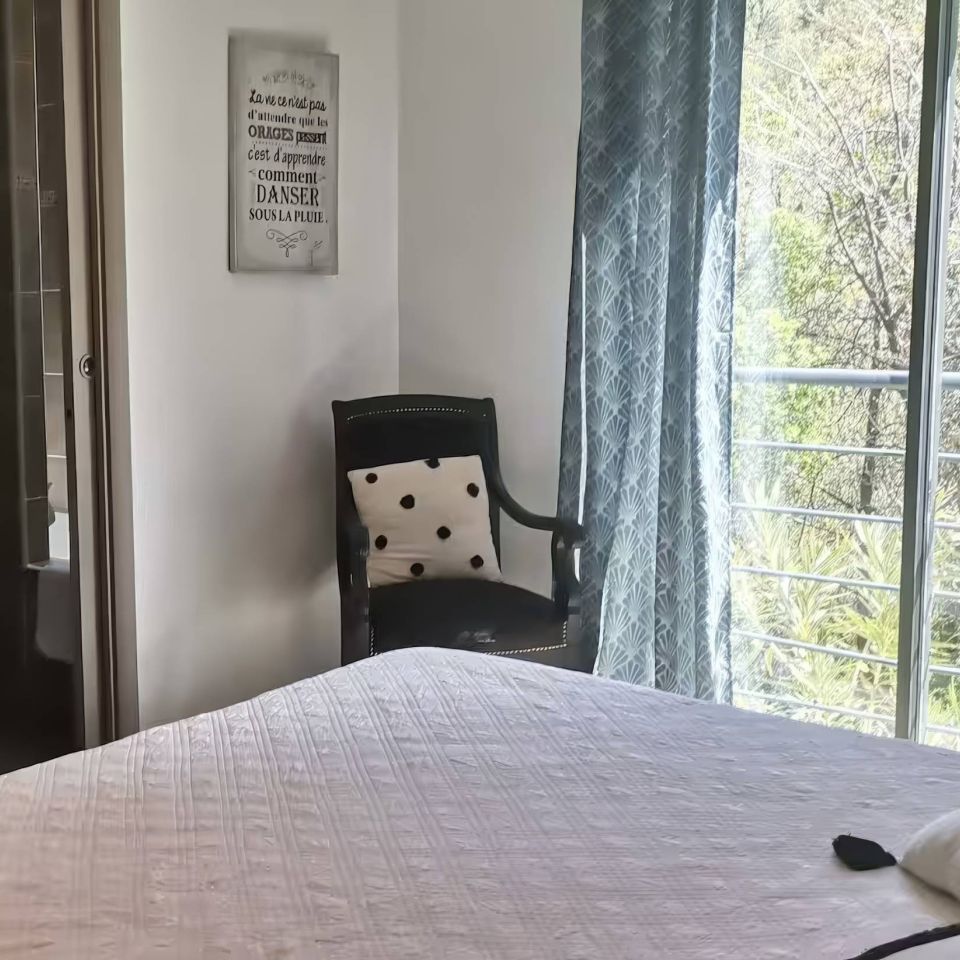 A bright, airy bedroom with a bed, a chair, patterned curtains, and a view of greenery outside.