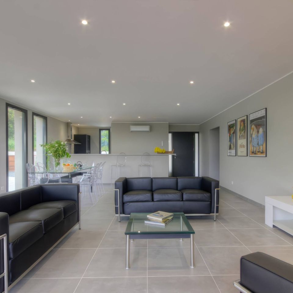 Modern lounge with black sofas, glass table, and an open kitchen area, featuring bright lighting and large windows.