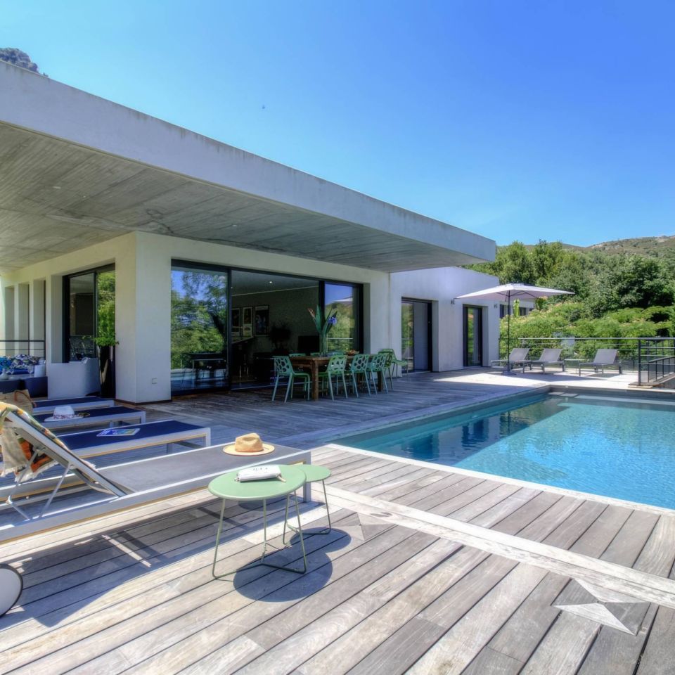 Modern house with flat roof, pool, and outdoor seating surrounded by greenery on a bright, sunny day.